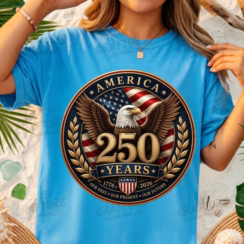 K&ouml;nnte beinhalten: Ein hellblaues T-Shirt mit einer kreisf&ouml;rmigen Grafik, die einen Wei&szlig;kopfseeadler, die amerikanische Flagge und den Text AMERICA 250 YEARS 1776-2026 OUR PAST OUR FUTURE zeigt. Das Design enth&auml;lt goldene Lorbeerzweige und ein gestreiftes Wappen.