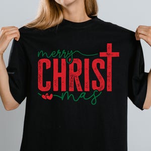Puede incluir: Camiseta negra con las palabras "Merry CHRISTmas" en rojo y verde. La palabra "CHRIST" est&aacute; en negrita, con un efecto desgastado, y una cruz reemplaza la letra "T". La palabra "merry" est&aacute; escrita en cursiva.
