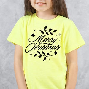 May include: A light yellow t-shirt with the words "Merry Christmas" in a black script font, surrounded by black leaves, stars, and dots. The shirt is short-sleeved and suitable for children.