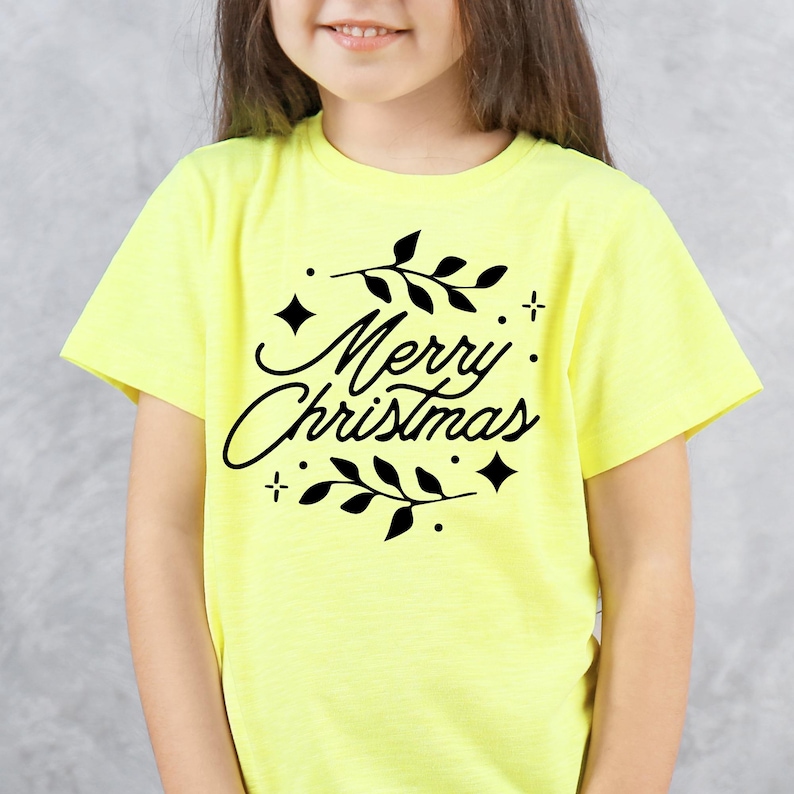 May include: A light yellow t-shirt with the words "Merry Christmas" in a black script font, surrounded by black leaves, stars, and dots. The shirt is short-sleeved and suitable for children.