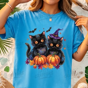 May include: A bright blue t-shirt featuring a graphic of two black kittens with large orange eyes. One kitten wears a purple witch hat. Both sit behind orange pumpkins surrounded by colourful autumn leaves and two small black bats in the air.