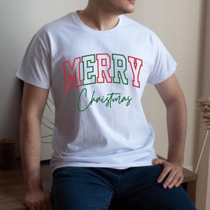 Puede incluir: Camiseta blanca con la frase "MERRY Christmas" en rojo y verde. La palabra "MERRY" est&aacute; en un estilo de letras de bloque con contorno rojo y relleno verde. "Christmas" est&aacute; escrito en cursiva verde.