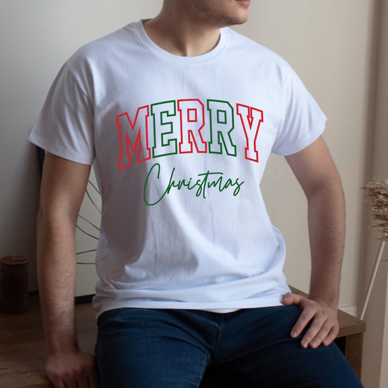 Puede incluir: Camiseta blanca con la frase "MERRY Christmas" en rojo y verde. La palabra "MERRY" est&aacute; en un estilo de letras de bloque con contorno rojo y relleno verde. "Christmas" est&aacute; escrito en cursiva verde.