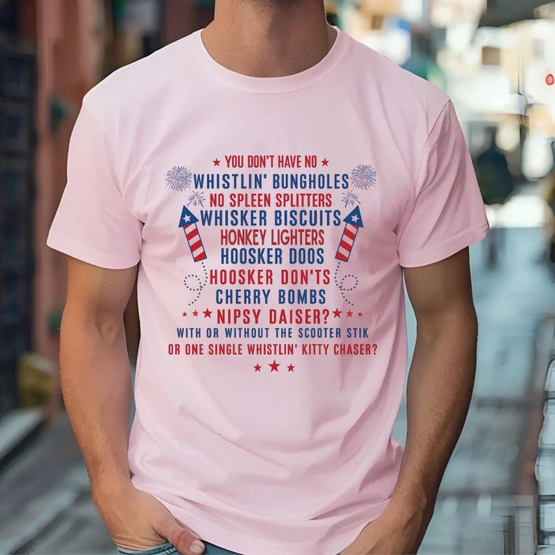 Op de afbeelding: Een lichtroze t-shirt met een patriottisch ontwerp. Het shirt heeft tekst in rood en blauw, waaronder zinnen als "Whistlin' Bungholes" en "Cherry Bombs", samen met ster- en vuurwerkgraphics. Het t-shirt is een casual kledingstuk.