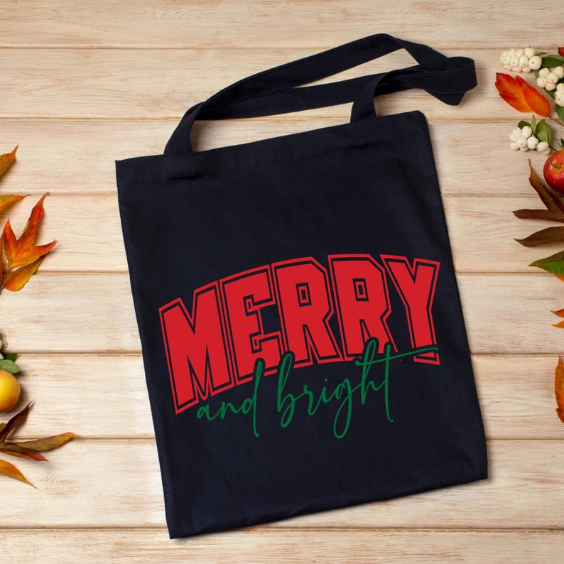 Puede incluir: Un bolso tote de color azul marino con asas negras. La bolsa presenta las palabras "MERRY" en letras rojas con un contorno rojo, y "and bright" en verde. La bolsa est&aacute; hecha de una tela resistente y es adecuada para transportar art&iacute;culos.