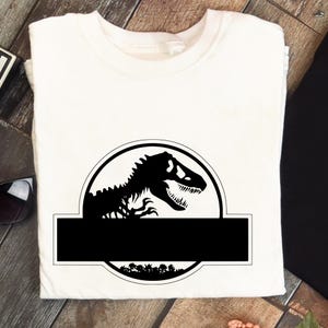 May include: A white t-shirt featuring a black graphic of a Tyrannosaurus Rex inside a circular emblem. Below the emblem is a black rectangle. The design includes a forest silhouette at the bottom of the circle.