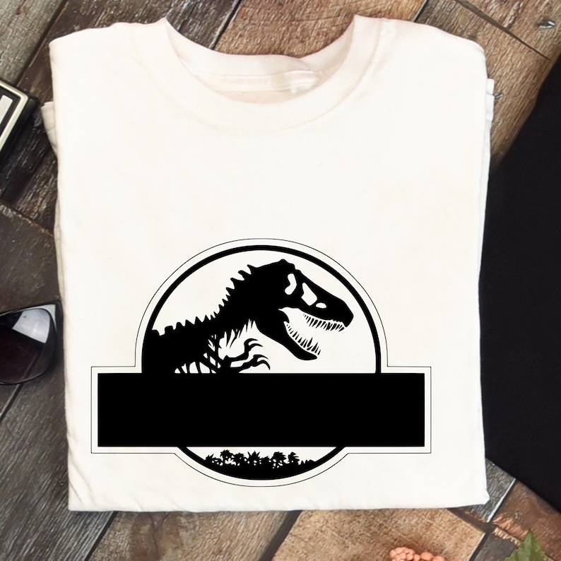 May include: A white t-shirt featuring a black graphic of a Tyrannosaurus Rex inside a circular emblem. Below the emblem is a black rectangle. The design includes a forest silhouette at the bottom of the circle.