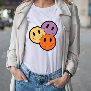 May include: White t-shirt with a graphic of three smiling faces in yellow, orange, and purple, each with a black outline. The shirt is worn with blue jeans and a light-coloured jacket.