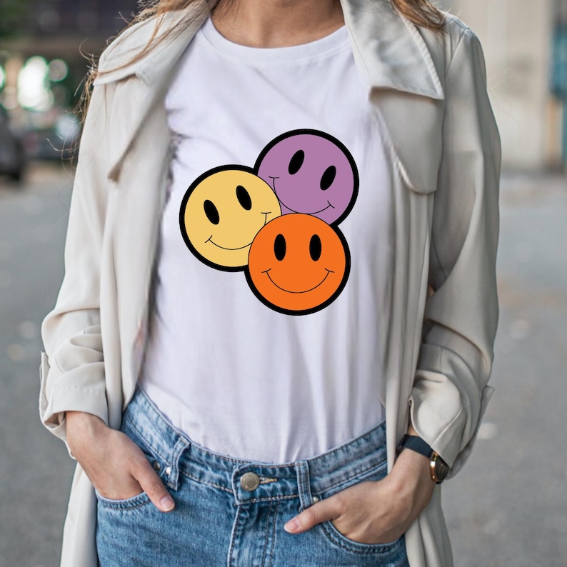 May include: White t-shirt with a graphic of three smiling faces in yellow, orange, and purple, each with a black outline. The shirt is worn with blue jeans and a light-coloured jacket.
