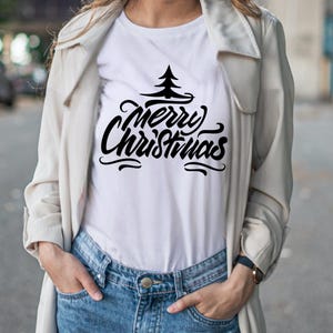 May include: White t-shirt with the words "Merry Christmas" in a black, stylized font, topped with a simple tree graphic. The shirt is paired with blue jeans and a light-colored coat.