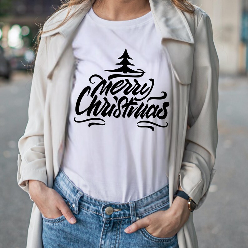 May include: White t-shirt with the words "Merry Christmas" in a black, stylized font, topped with a simple tree graphic. The shirt is paired with blue jeans and a light-colored coat.
