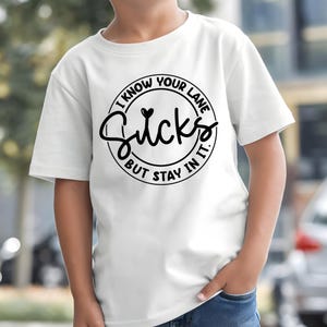 K&ouml;nnte beinhalten: Wei&szlig;es T-Shirt mit einem schwarzen kreisf&ouml;rmigen Aufdruck mit der Aufschrift "I KNOW YOUR LANE Sucks BUT STAY IN IT." Das Shirt hat kurze &Auml;rmel und wird von einer Person getragen.