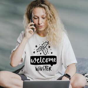 May include: A white t-shirt with the words "welcome WINTER" in black, along with a holly sprig design. The shirt is being worn by a person with long, curly blonde hair, who is on a phone call.