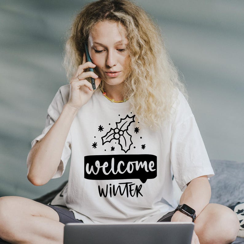 May include: A white t-shirt with the words "welcome WINTER" in black, along with a holly sprig design. The shirt is being worn by a person with long, curly blonde hair, who is on a phone call.