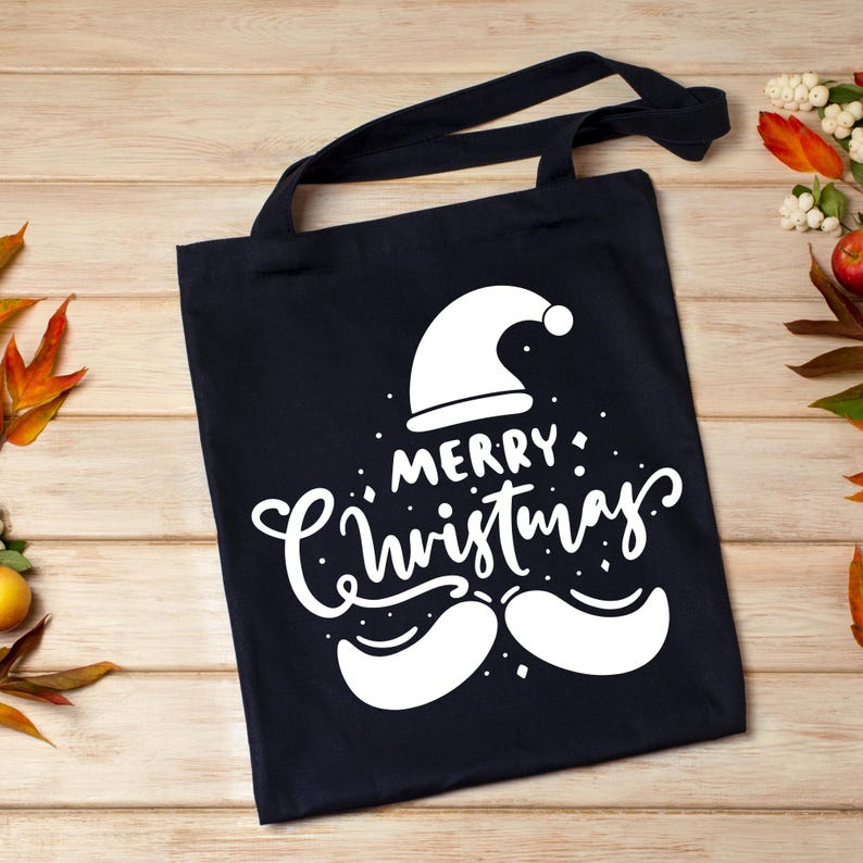 May include: A navy blue tote bag with black handles. The bag features a white graphic of a Santa hat, mustache, and the words "MERRY Christmas" in a whimsical font. The bag is made of a sturdy fabric.