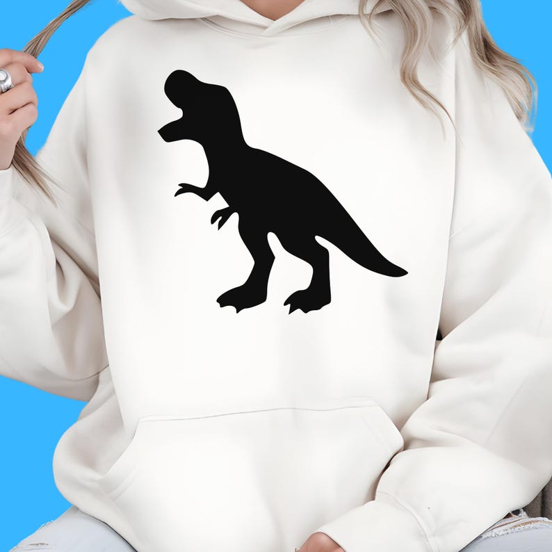 May include: Cream-colored hoodie featuring a black Tyrannosaurus Rex silhouette on the front. The dinosaur is depicted in a side profile with its mouth open. The hoodie has a kangaroo pocket and a drawstring hood.