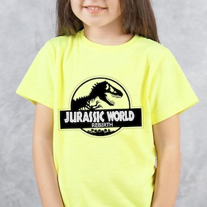May include: A yellow t-shirt featuring the "Jurassic World" logo in black and white. The logo includes a dinosaur silhouette inside a circle, with the text "JURASSIC WORLD" and "REBIRTH" below.