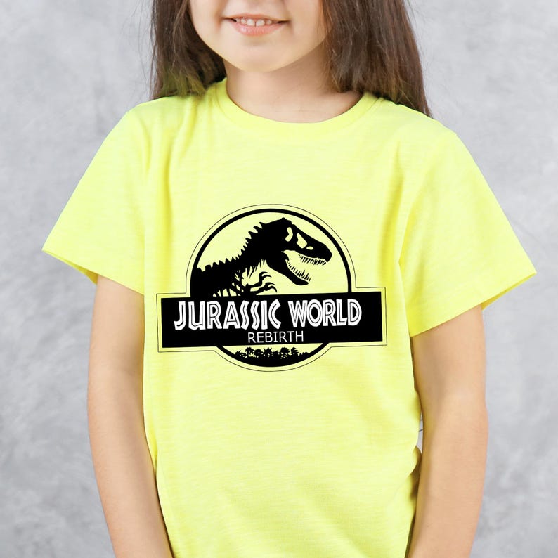 May include: A yellow t-shirt featuring the "Jurassic World" logo in black and white. The logo includes a dinosaur silhouette inside a circle, with the text "JURASSIC WORLD" and "REBIRTH" below.
