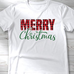 Puede incluir: Camiseta blanca con las palabras "MERRY" en letras a cuadros rojas y negras sobre la palabra "Christmas" en escritura verde. Una prenda casual y festiva.