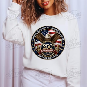 K&ouml;nnte beinhalten: Ein wei&szlig;es Langarm-Sweatshirt mit einer kreisf&ouml;rmigen Grafik, die einen Wei&szlig;kopfseeadler, eine amerikanische Flagge und den Text 250th ANNIVERSARY 1776-2026, HONORING OUR PAST, CELEBRATING OUR PRESENT, INSPIRING OUR FUTURE, umgeben von goldenen Sternen zeigt.