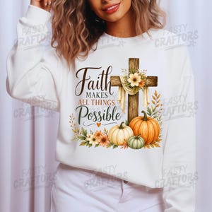 May include: A white long-sleeve sweatshirt featuring a graphic print of a wooden cross draped with fabric and flowers, accompanied by three pumpkins and autumn foliage. The text reads Faith MAKES ALL THINGS Possible with a small orange heart.