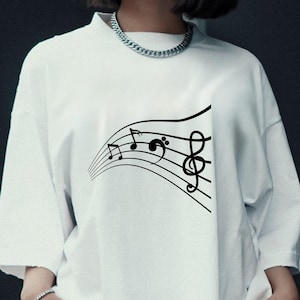 May include: White t-shirt featuring a black musical staff design with treble and bass clef symbols and musical notes. The graphic is a flowing, curved design. The shirt has a crew neck and short sleeves.