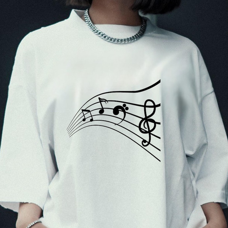 May include: White t-shirt featuring a black musical staff design with treble and bass clef symbols and musical notes. The graphic is a flowing, curved design. The shirt has a crew neck and short sleeves.