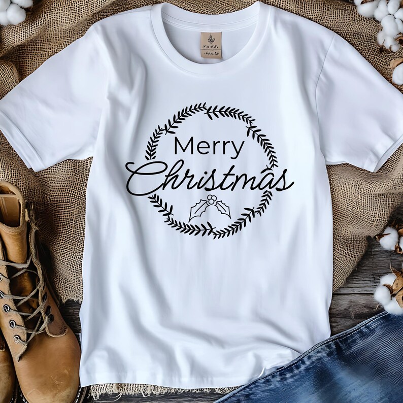 May include: White t-shirt with a black wreath design and the words "Merry Christmas" in a script font. The wreath surrounds the text and includes holly leaves. The shirt is displayed on a rustic background.
