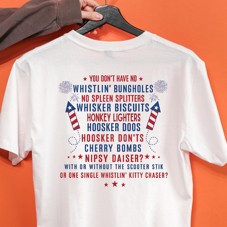 Op de afbeelding: Wit T-shirt met rode en blauwe tekst en vuurwerkgraphics. De tekst bevat zinnen als "Whistlin' Bungholes" en "Whisker Biscuits". Het T-shirt wordt door een persoon omhoog gehouden.