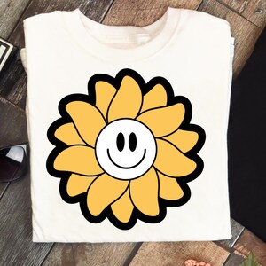 May include: Cream-coloured t-shirt with a large, stylised yellow flower design. The flower has a black outline and a white centre with a smiling face. The design is centrally placed, giving a cheerful and playful look.