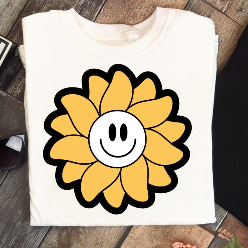 May include: Cream-coloured t-shirt with a large, stylised yellow flower design. The flower has a black outline and a white centre with a smiling face. The design is centrally placed, giving a cheerful and playful look.