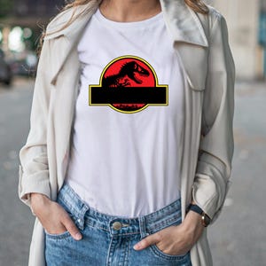 May include: White t-shirt featuring a black dinosaur silhouette inside a red circle with a yellow border. Below the circle is a black rectangle. The shirt is worn by a person wearing blue jeans and a light-colored jacket.