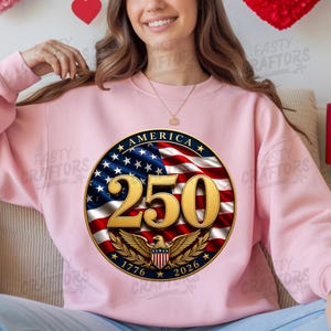 K&ouml;nnte beinhalten: Ein hellrosa Rundhals-Sweatshirt mit einer kreisf&ouml;rmigen Grafik auf dem Hintergrund der amerikanischen Flagge. Das Design zeigt den goldenen Text 250, das Wort AMERICA und die Jahreszahlen 1776 und 2026 mit einem goldenen Adleremblem.