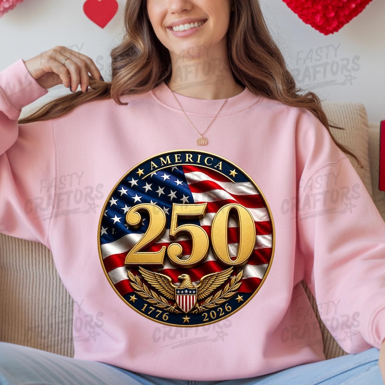 K&ouml;nnte beinhalten: Ein hellrosa Rundhals-Sweatshirt mit einer kreisf&ouml;rmigen Grafik auf dem Hintergrund der amerikanischen Flagge. Das Design zeigt den goldenen Text 250, das Wort AMERICA und die Jahreszahlen 1776 und 2026 mit einem goldenen Adleremblem.