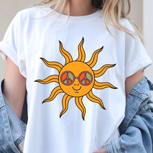 May include: White t-shirt with a cartoon sun design. The sun has orange rays, a smiling face, and wears round glasses with peace signs. The design is centrally placed, giving a retro and cheerful appearance.