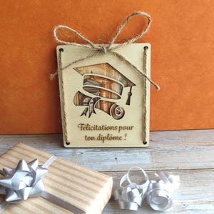 Peut inclure: Une plaque rectangulaire en bois clair avec une découpe en forme de toque de diplômé et de diplôme. La découpe révèle une image imprimée de colonnes architecturales. Le texte "Félicitations pour ton diplôme !" est gravé en dessous. Un nœud en ficelle de jute est attaché en haut.