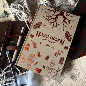 Puede incluir: Un libro titulado "HAZELTHORN" de C.G. Drews, con una cubierta beige con un diseño de rama de árbol marrón, mariposas e ilustraciones botánicas. El libro está rodeado de materiales de arte y otros libros.