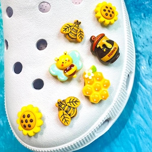 May include: A white clog shoe decorated with six colourful charms featuring a yellow sunflower, a brown honey pot with the text Honey, a yellow honeycomb, and three different bee designs in yellow, black, and light blue colours attached to the surface.