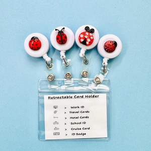 May include: Four white retractable badge reels featuring ladybug embellishments attached to a clear plastic card holder. The card holder displays the text Retractable Card Holder followed by a list of uses including Work ID, Travel Cards, Hotel Cards, School ID, Cruise Card, and ID Badge.