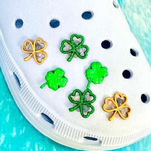 Può includere: Scarpa bianca con diversi ciondoli a forma di trifoglio verde e oro. I ciondoli sono in vari stili, tra cui verde glitterato e tinta unita, e oro con centri trasparenti. La scarpa ha diversi fori rotondi.