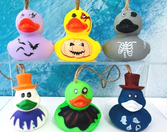 Könnte beinhalten: Sechs Gummi-Enten-Ornamente, die an einer Schnur hängen. Die obere Reihe zeigt eine lavendelfarbene Ente mit Fledermäusen, eine gelbe mit Kürbis und eine graue mit Skelettaufdruck. Die untere Reihe zeigt eine weiße Ente mit Zylinder, eine grüne mit Umhang und eine blaue mit Zylinder und Geistermotiven.