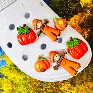May include: A white foam clog shoe decorated with four charms featuring two orange pumpkins with green stems and two scarecrows wearing orange overalls and hats. The charms are arranged on the surface of the shoe, which has circular ventilation holes.