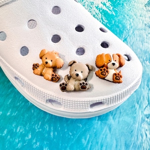 May include: Three small, molded plastic dog-shaped charms attached to the top of a white foam clog. The charms feature a light brown puppy, a grey puppy, and a brown and white puppy, each depicted in a sitting position with visible paw details.