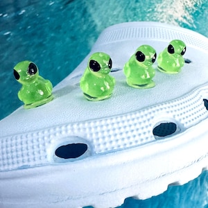 May include: Four small, translucent neon green duck-shaped charms with solid black eyes are attached to the top surface of a white foam clog shoe. The shoe features a textured, perforated surface with circular ventilation holes along the side.