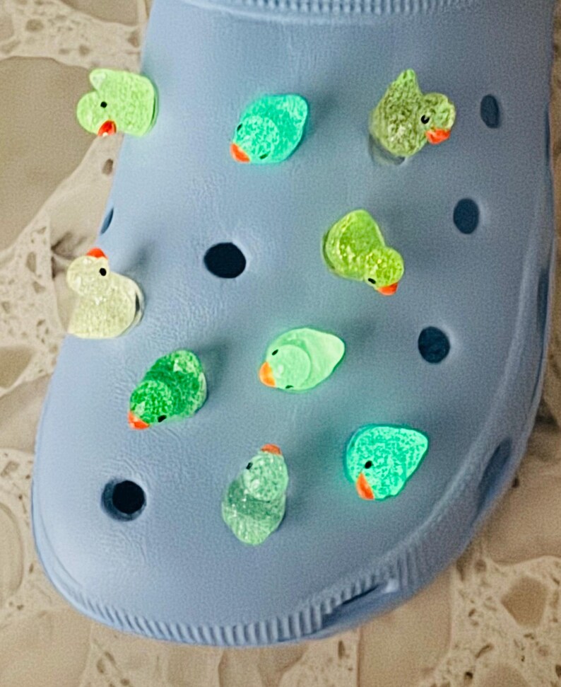 May include: A light blue clog shoe adorned with multiple green duck-shaped charms. The charms have orange beaks and black eyes. The shoe is made of a rubbery material and has several ventilation holes.