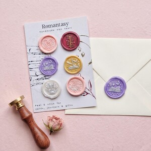 May include: A selection of handmade wax seals in shades of pink, purple, and gold, featuring detailed designs. The seals are presented on a card with the text "Romantasy" and a wooden handled wax stamp. A dried rose and an envelope are also visible.
