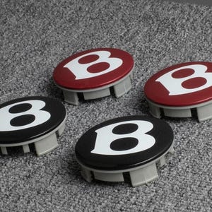 Op de afbeelding: Vier wieldopjes. Twee zijn zwart met een wit "B"-logo en twee zijn rood met een wit "B"-logo. De doppen hebben een grijze basis en staan tegen een grijze, getextureerde achtergrond.