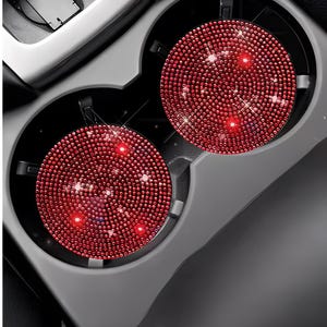 May include: Two red, circular car cup holder coasters. Each coaster is covered in small, sparkling red rhinestones. The coasters are placed in a gray car console cup holder. The image is taken from a top-down perspective.
