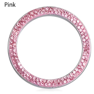 Puede incluir: Un anillo decorativo circular rosa con un marco de metal plateado. El anillo está incrustado con numerosos cristales rosas pequeños y brillantes. La palabra "Pink" está impresa en la esquina superior izquierda.