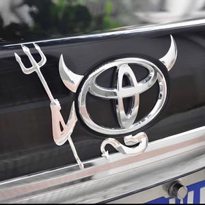 May include: A chrome-finished car emblem featuring the Toyota logo with devil horns and a trident. The emblem is mounted on a black vehicle surface with a chrome trim. The design adds a unique, edgy aesthetic to the car.